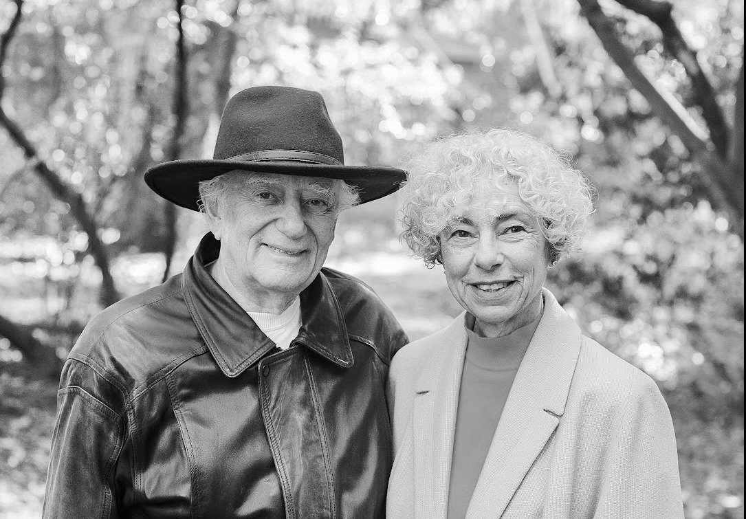 Black-and-white photograph of David Rosenthal, standing on the left in a dark leather jacket and fedora, and Vicky Reich, standing on the right in a light-colored blazer and mock turtleneck top.